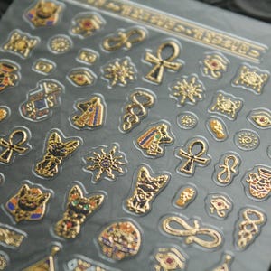 May include: A sheet of gold and black Egyptian themed stickers. The stickers feature ankh symbols, scarab beetles, eyes of Horus, and other Egyptian motifs.