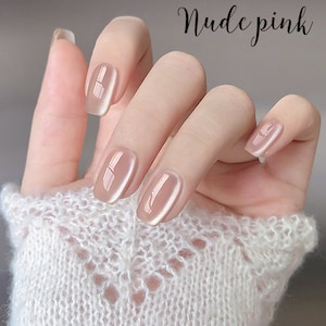 Puede incluir: Una mano con u&ntilde;as de gel rosa nude. Las u&ntilde;as son de un rosa suave y transl&uacute;cido con un acabado brillante. El texto "Nude pink" est&aacute; escrito en cursiva. La mano lleva un su&eacute;ter blanco de punto abierto.