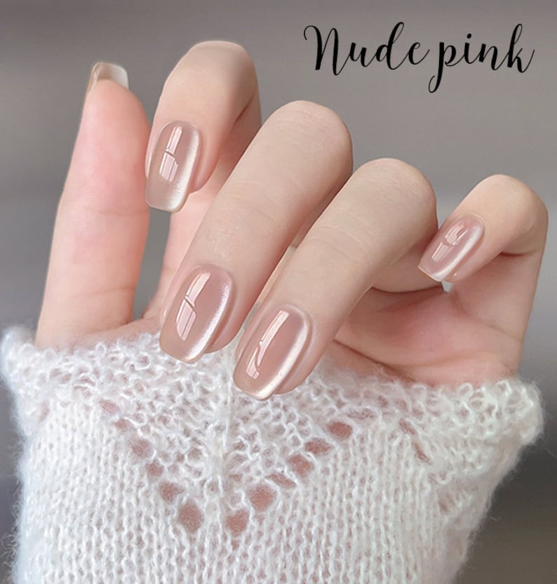 Puede incluir: Una mano con u&ntilde;as de gel rosa nude. Las u&ntilde;as son de un rosa suave y transl&uacute;cido con un acabado brillante. El texto "Nude pink" est&aacute; escrito en cursiva. La mano lleva un su&eacute;ter blanco de punto abierto.