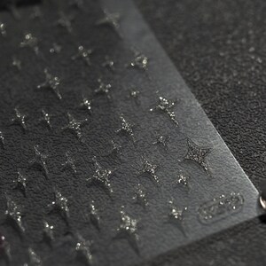 May include: A sheet of clear nail art stickers with a repeating pattern of silver glitter stars. The sheet is on a black textured surface.