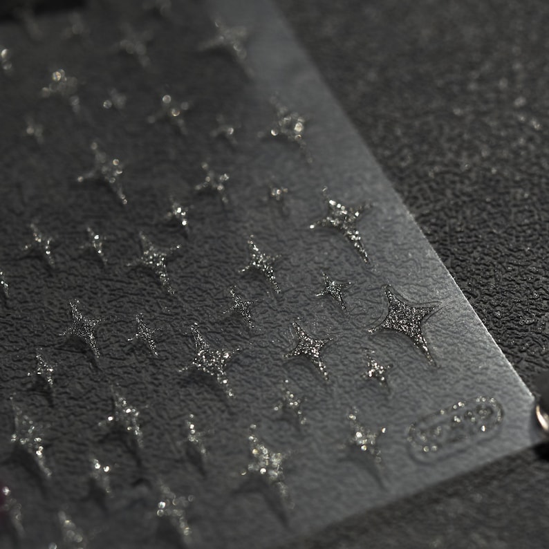 May include: A sheet of clear nail art stickers with a repeating pattern of silver glitter stars. The sheet is on a black textured surface.