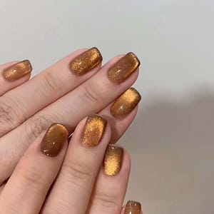 May include: Fingernails featuring a cat-eye polish effect in a deep, metallic bronze shade. The finish is highly reflective with a shimmering, glittery texture that creates a bright, concentrated band of light across the center of each nail.