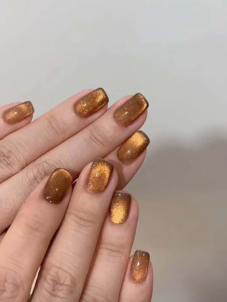 May include: Fingernails featuring a cat-eye polish effect in a deep, metallic bronze shade. The finish is highly reflective with a shimmering, glittery texture that creates a bright, concentrated band of light across the center of each nail.