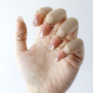 Puede incluir: Una mano con uñas largas y ovaladas adornadas con anillos de uñas dorados. Las uñas tienen un esmalte rosa claro brillante. Los anillos son delicados y envuelven la base de las uñas, añadiendo un toque de elegancia.