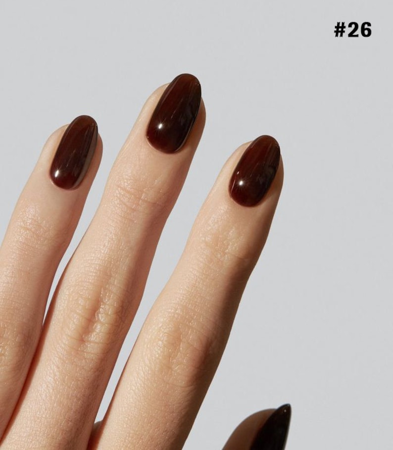 May include: Three fingernails shaped in an oval style, each coated in a glossy, deep dark brown nail polish. The nails are displayed against a neutral background, with the text #26 visible in the upper right corner of the frame.