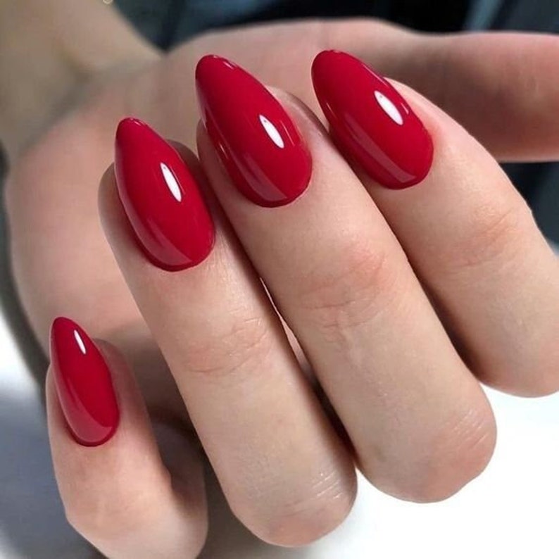 May include: A set of four fingernails shaped into a pointed almond style, each coated in a vibrant, glossy, solid red nail polish. The finish is smooth and reflective, highlighting the bright crimson pigment across the entire nail surface.