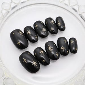 Puede incluir: Un juego de diez uñas postizas negras brillantes, cada una con un diseño celestial de estrellas doradas y pequeños puntos. Las uñas están dispuestas en dos filas, mostrando un acabado liso y reflectante con patrones de estrellas puntiagudas.