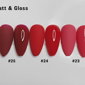 May include: Six almond-shaped nail tips arranged in pairs of matte and glossy finishes in shades of red. The text "Matt & Gloss" appears at the top, with labels "#25", "#24", and "#23" positioned below the corresponding pairs of nail samples.