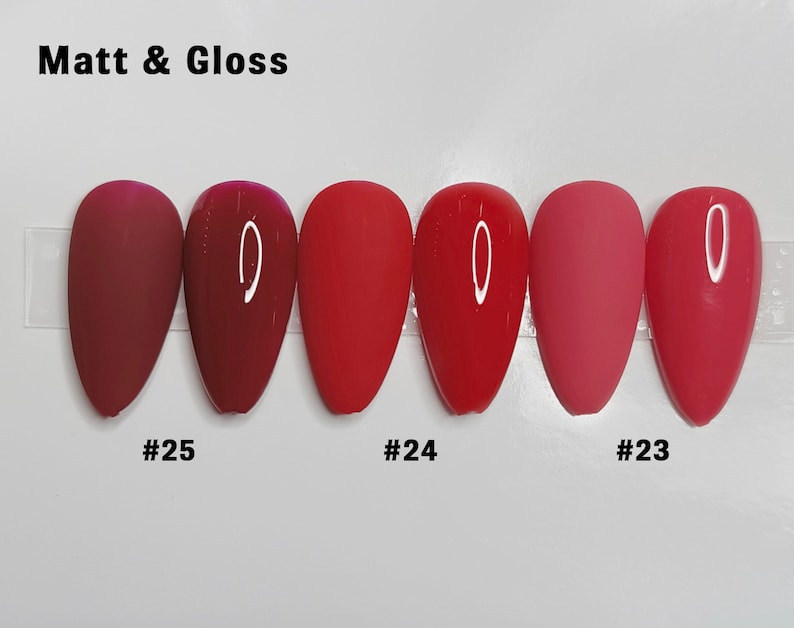 May include: Six almond-shaped nail tips arranged in pairs of matte and glossy finishes in shades of red. The text "Matt & Gloss" appears at the top, with labels "#25", "#24", and "#23" positioned below the corresponding pairs of nail samples.