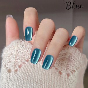 Puede incluir: Primer plano de una mano con esmalte de u&ntilde;as azul verdoso brillante. Las u&ntilde;as son de forma ovalada. La palabra "Blue" est&aacute; escrita en cursiva. La mano lleva un su&eacute;ter blanco tejido.