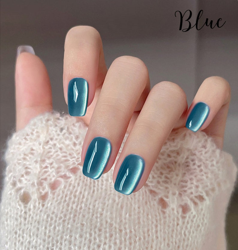 Puede incluir: Primer plano de una mano con esmalte de u&ntilde;as azul verdoso brillante. Las u&ntilde;as son de forma ovalada. La palabra "Blue" est&aacute; escrita en cursiva. La mano lleva un su&eacute;ter blanco tejido.