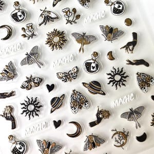 May include: A sheet of clear stickers with a variety of black and gold designs, including moons, stars, butterflies, planets, hands, and the word "MAGIC".