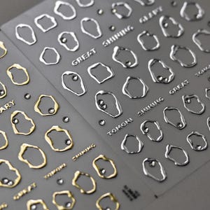 Könnte beinhalten: Nahaufnahme von zwei Bögen Nagelkunst-Aufklebern. Ein Bogen zeigt goldfarbene, unregelmäßig geformte Umrisse mit kleinen, silbernen Verzierungen. Der andere Bogen hat silberfarbene Umrisse. Die Wörter "GREAT SHINING" und "TOKORI SHINING" sind aufgedruckt.