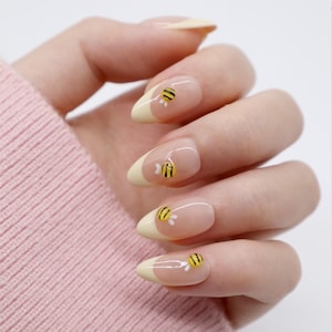 May include: Oval-shaped fingernails featuring a light yellow French tip manicure. Each nail is decorated with a small, three-dimensional yellow and black striped bee charm with tiny white wings positioned near the base of the yellow tip.