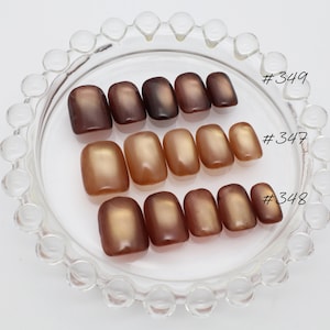 May include: Three sets of press-on nails arranged on a clear glass dish with a beaded rim. The sets are labeled #349, #347, and #348. Each set features a smooth, glossy finish with a cat-eye effect in varying shades of brown, bronze, and copper.