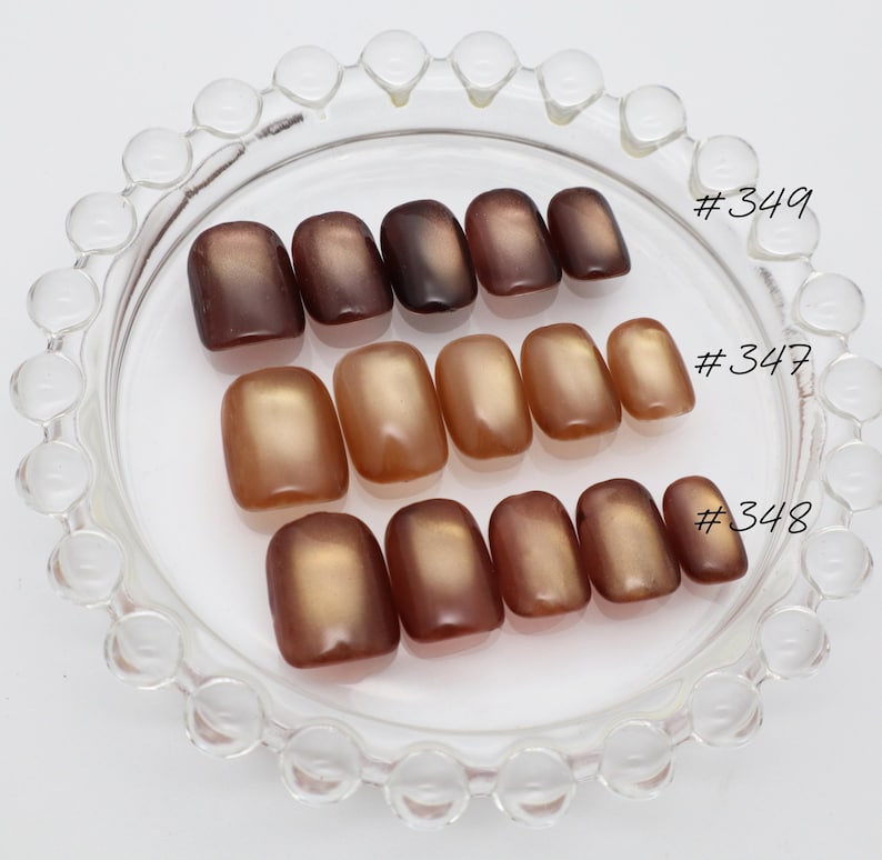 May include: Three sets of press-on nails arranged on a clear glass dish with a beaded rim. The sets are labeled #349, #347, and #348. Each set features a smooth, glossy finish with a cat-eye effect in varying shades of brown, bronze, and copper.
