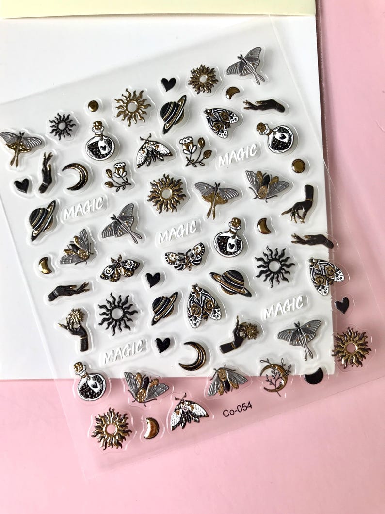 May include: A sheet of clear sticker paper with gold and black illustrations of moons, stars, planets, butterflies, and hands. The word "MAGIC" is repeated several times on the sheet. The sheet is labeled "CO-054".