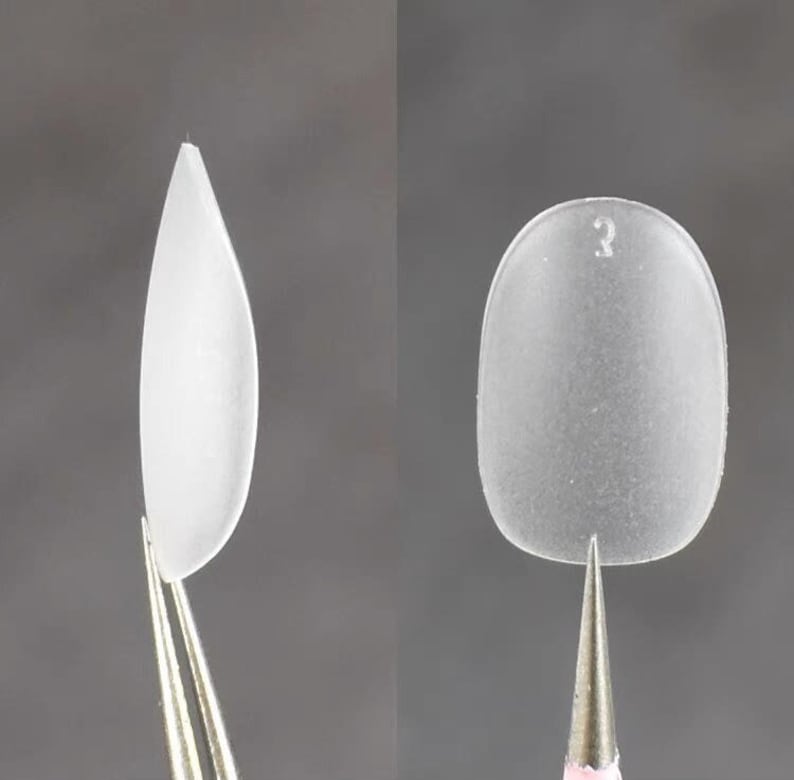 May include: Two clear, frosted artificial nail tips held by metal tweezers. The left tip is almond-shaped and pointed. The right tip is rounded and features the number 3 embossed near the top edge. Both items have a smooth, matte finish.