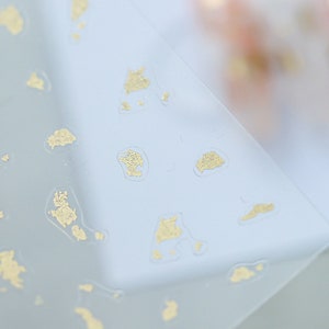 May include: A sheet of clear plastic with gold foil accents. The foil is in various shapes and sizes, creating a speckled pattern.