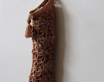 May include: A hanging brown crochet bag with a decorative tassel and wooden bead. The bag has a net-like design and is suspended against a white wall. The bag is designed to hold items.