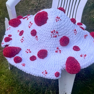 May include: A white crocheted mushroom hat with red, textured spots and small red bead accents. The hat is sitting on a white chair, showcasing its unique, whimsical design. This handmade hat is a fun accessory.