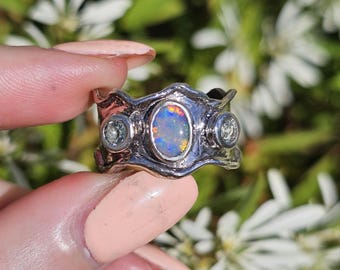 May include: A silver ring featuring an oval opal centre stone and two smaller round stones. The opal exhibits a vibrant play of colour. The ring has a textured, wavy design.