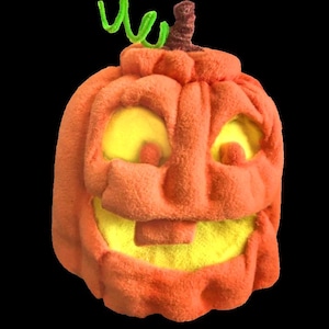 May include: A plush pumpkin toy with a soft orange exterior and a bright yellow interior visible through the carved-out eyes and mouth. The pumpkin features a brown stem and a curly, bright green pipe cleaner vine attached to the top.