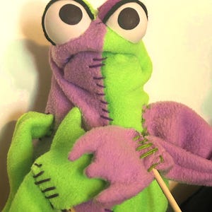 Könnte beinhalten: Eine Handpuppe mit einem zweifarbigen Design aus hellgrünem und lavendelfarbenem Fleecestoff. Die Puppe hat zwei große weiße Augen mit schwarzen Pupillen, sichtbare dunkelviolette Nähte entlang der Mittelnaht und zwei hölzerne Steuerstäbe.