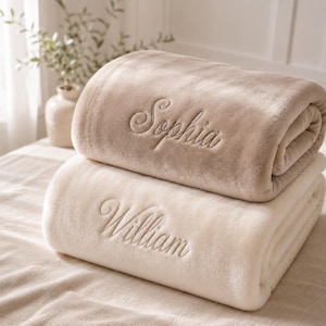 May include: Two soft blankets, one in a beige hue and the other cream-coloured, are stacked. The top blanket is embroidered with the name "Sophia" in a cursive script. The bottom blanket features the name "William".