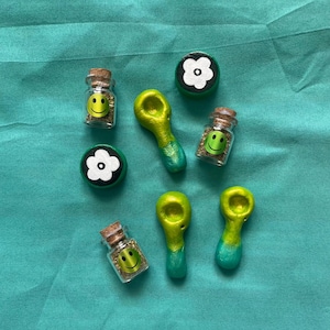 Pode incluir: Uma variedade de pequenos frascos de vidro com rolhas, pequenos cachimbos verdes e amarelos e objetos em forma de flor em preto e branco. Os frascos cont&ecirc;m rostos sorridentes amarelos e mat&eacute;ria vegetal verde.