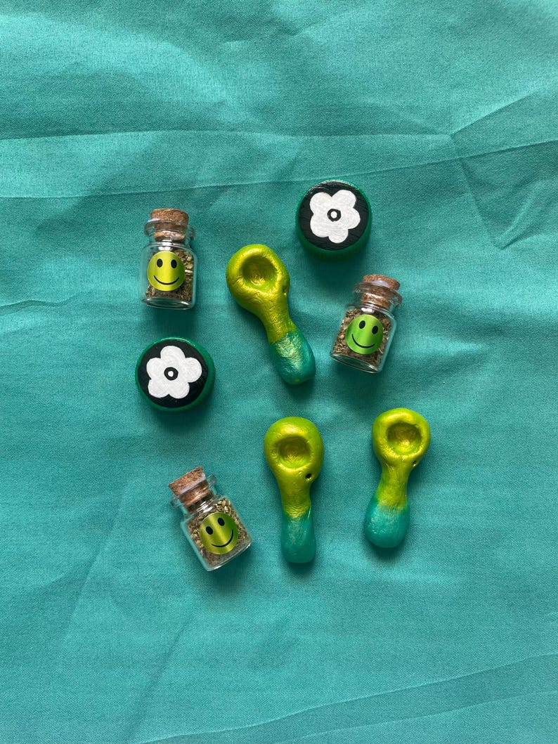 Pode incluir: Uma variedade de pequenos frascos de vidro com rolhas, pequenos cachimbos verdes e amarelos e objetos em forma de flor em preto e branco. Os frascos cont&ecirc;m rostos sorridentes amarelos e mat&eacute;ria vegetal verde.