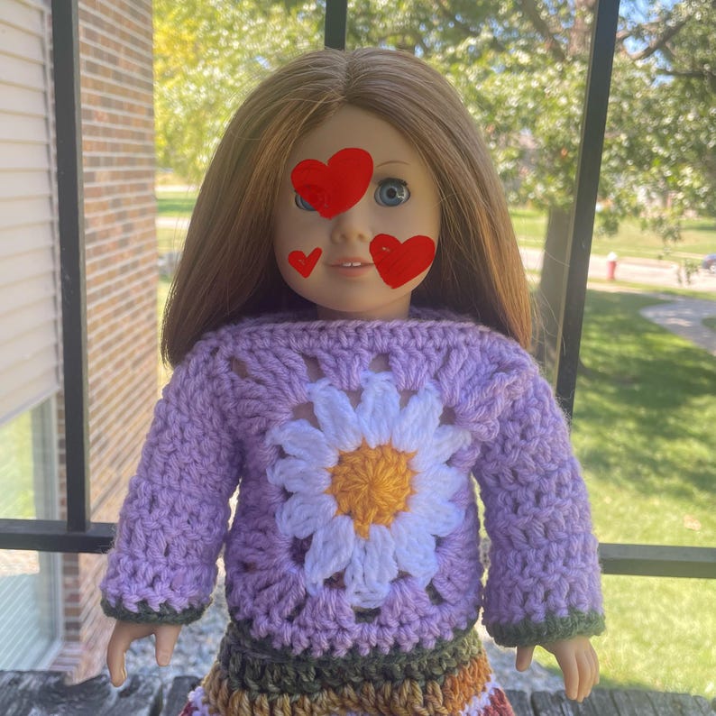K&ouml;nnte beinhalten: Ein lilafarbener H&auml;kelpullover mit einem wei&szlig;en G&auml;nsebl&uuml;mchenmuster. Der Pullover wird von einer Puppe mit braunen Haaren und blauen Augen getragen.