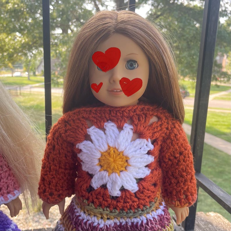 K&ouml;nnte beinhalten: Eine Puppe tr&auml;gt einen geh&auml;kelten orangen Pullover mit einem wei&szlig;en G&auml;nsebl&uuml;mchen-Design und einen mehrfarbigen geh&auml;kelten Rock.