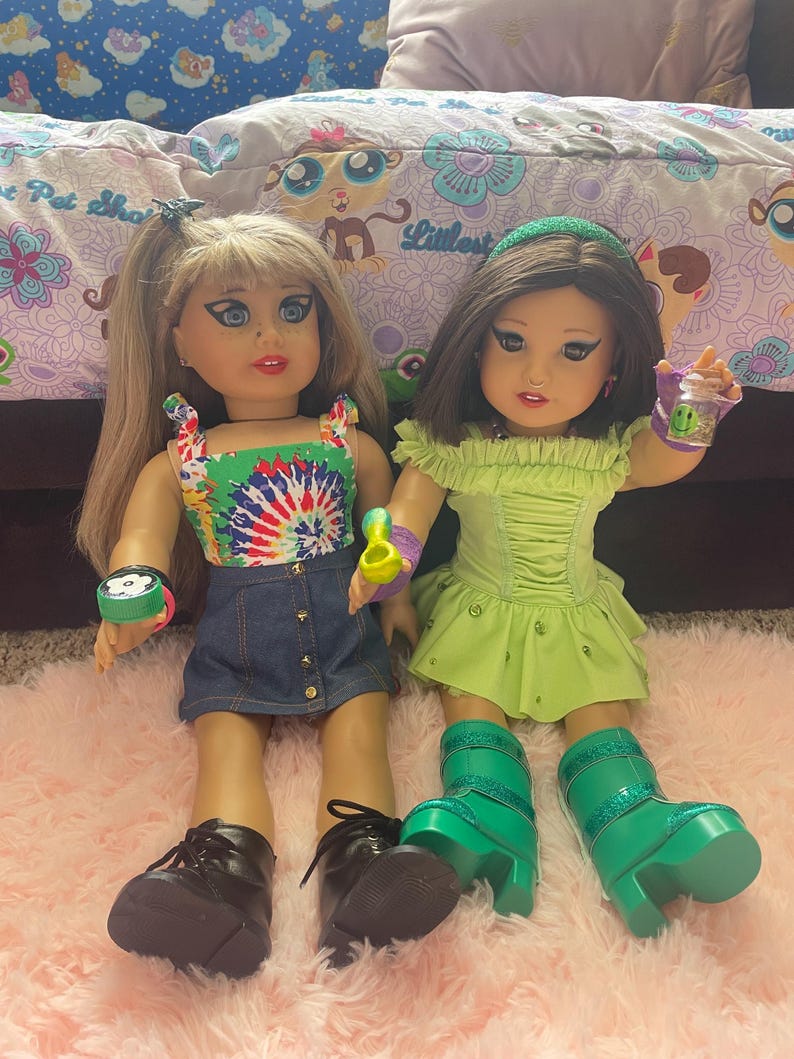 Pode incluir: Duas bonecas s&atilde;o exibidas, cada uma com cabelos longos e tra&ccedil;os faciais detalhados. Uma boneca usa um top tie-dye, uma saia jeans e sapatos pretos. A outra boneca est&aacute; em um vestido e botas verdes. Os acess&oacute;rios incluem pulseiras e uma x&iacute;cara pequena.