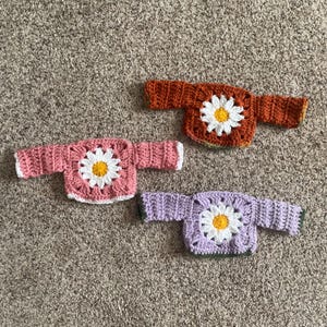 K&ouml;nnte beinhalten: Drei geh&auml;kelte Pullover mit G&auml;nsebl&uuml;mchen-Designs. Die Pullover sind in Rosa, Lila und Orange.