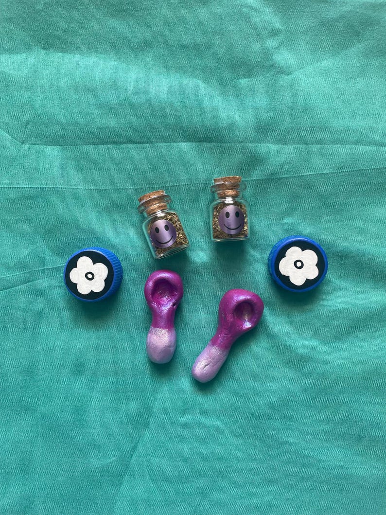Pode incluir: Uma variedade de itens decorativos. Duas pequenas garrafas de vidro com rolhas de corti&ccedil;a, cada uma contendo glitter dourado e um rosto sorridente. Duas tampas de garrafa azuis com desenhos de flores brancas e dois objetos roxos em forma de colher.