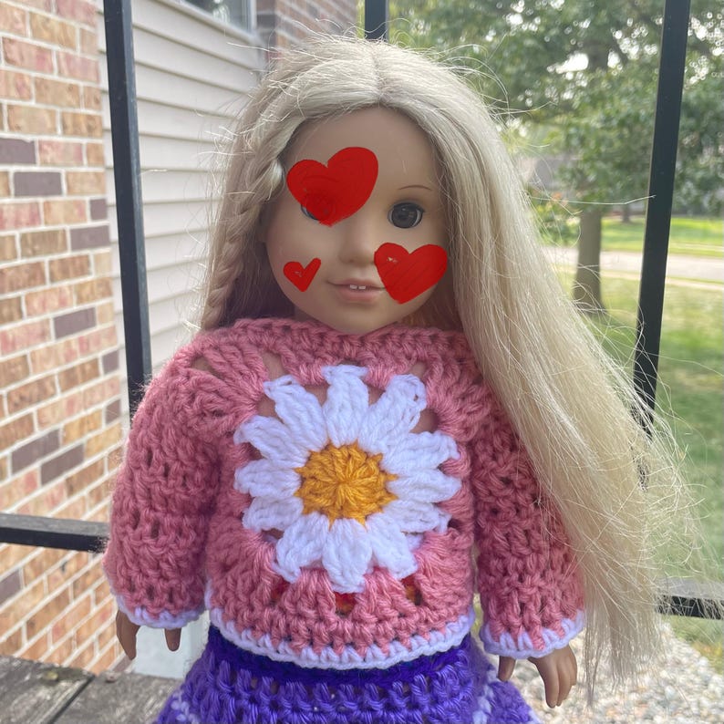 K&ouml;nnte beinhalten: Eine Puppe tr&auml;gt einen rosa geh&auml;kelten Pullover mit einem wei&szlig;en G&auml;nsebl&uuml;mchen-Design und einen lila geh&auml;kelten Rock.