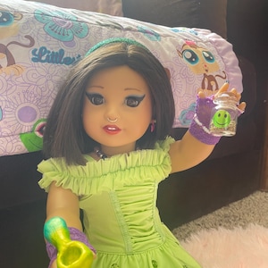 Pode incluir: Uma boneca vestindo um vestido verde lim&atilde;o com decote franzido e uma faixa de cabelo combinando. A boneca est&aacute; segurando um pequeno pote de vidro com um rosto sorridente e um cachimbo colorido. A boneca tem cabelo escuro e est&aacute; maquiada.