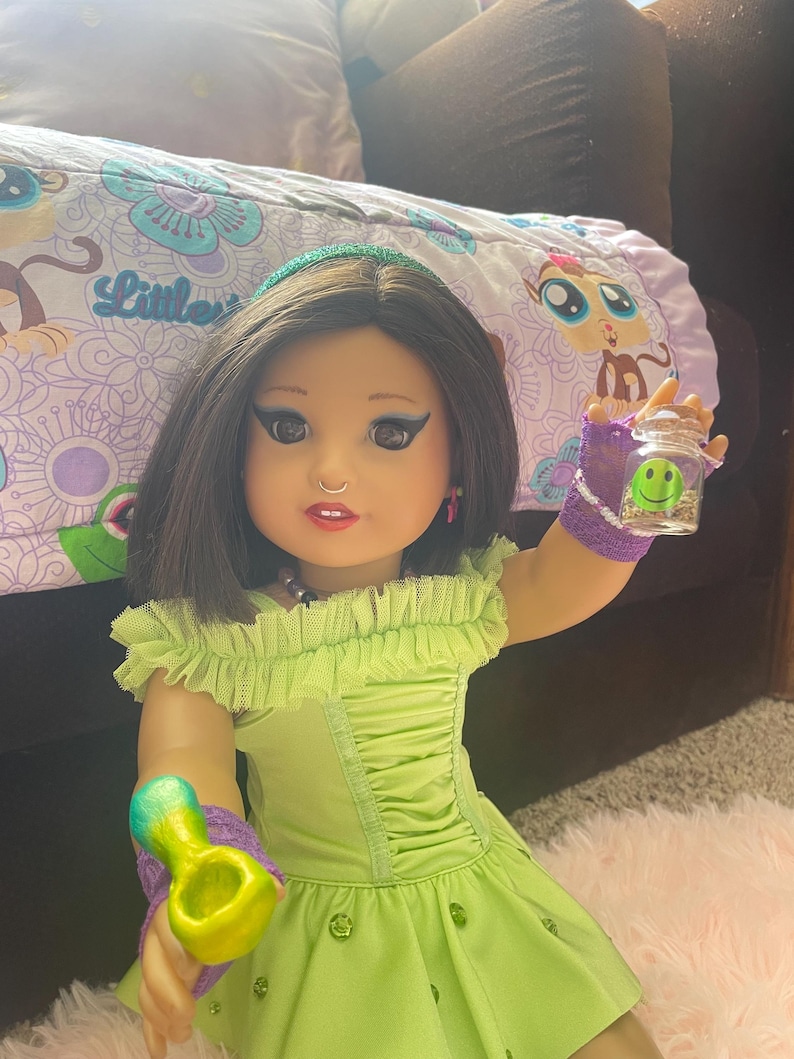 Pode incluir: Uma boneca vestindo um vestido verde lim&atilde;o com decote franzido e uma faixa de cabelo combinando. A boneca est&aacute; segurando um pequeno pote de vidro com um rosto sorridente e um cachimbo colorido. A boneca tem cabelo escuro e est&aacute; maquiada.