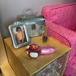 Pode incluir: Uma cena em miniatura com um cachimbo em forma de cogumelo vermelho e branco, uma pequena foto com moldura prateada, uma caixa prateada e uma garrafa de vidro com uma rolha. Os itens est&atilde;o dispostos em uma mesa de cabeceira de madeira com gavetas transparentes, ao lado de um sof&aacute; rosa estampado.