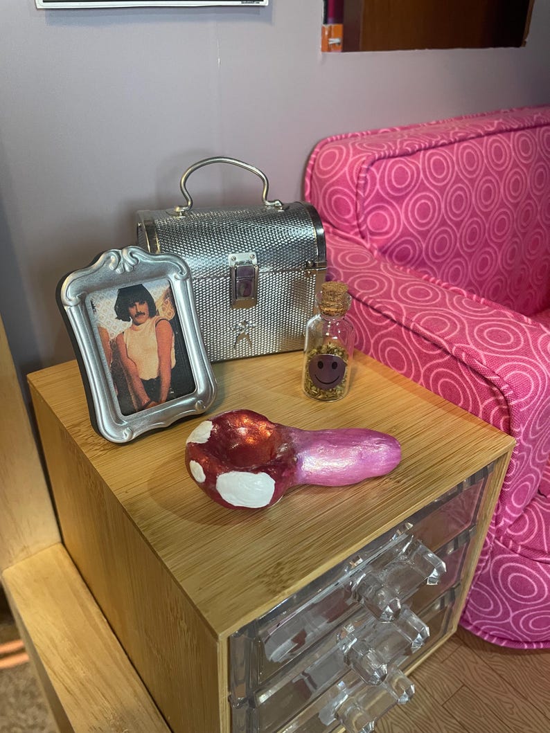 Pode incluir: Uma cena em miniatura com um cachimbo em forma de cogumelo vermelho e branco, uma pequena foto com moldura prateada, uma caixa prateada e uma garrafa de vidro com uma rolha. Os itens est&atilde;o dispostos em uma mesa de cabeceira de madeira com gavetas transparentes, ao lado de um sof&aacute; rosa estampado.