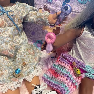 Pode incluir: Duas bonecas s&atilde;o mostradas, uma com um vestido floral e a outra com um top prateado. Elas est&atilde;o segurando pequenos cachimbos de vidro. Uma bolsa de croch&ecirc; com a palavra "believe" tamb&eacute;m &eacute; vis&iacute;vel, junto com uma garrafa pequena com um rosto sorridente.