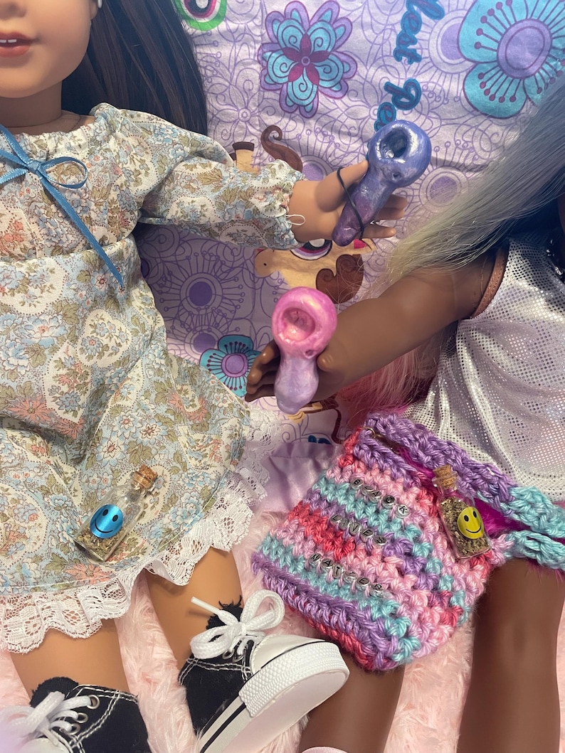 Pode incluir: Duas bonecas s&atilde;o mostradas, uma com um vestido floral e a outra com um top prateado. Elas est&atilde;o segurando pequenos cachimbos de vidro. Uma bolsa de croch&ecirc; com a palavra "believe" tamb&eacute;m &eacute; vis&iacute;vel, junto com uma garrafa pequena com um rosto sorridente.