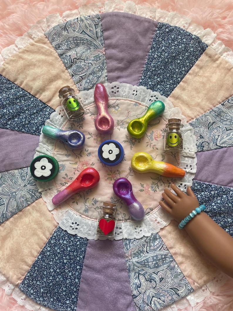 Pode incluir: Uma variedade de cachimbos pequenos pintados &agrave; m&atilde;o e potes decorativos com rolhas de corti&ccedil;a. Os cachimbos s&atilde;o de v&aacute;rias cores, incluindo rosa, verde e roxo. Os potes cont&ecirc;m rostos sorridentes e um cora&ccedil;&atilde;o vermelho. Dispostos sobre um edredom com estampa floral.