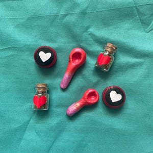 Pode incluir: Uma variedade de itens feitos &agrave; m&atilde;o, incluindo dois pequenos frascos de vidro com rolhas de corti&ccedil;a, cada um contendo um cora&ccedil;&atilde;o vermelho e ervas secas. Duas colheres vermelhas e rosa e dois c&iacute;rculos pretos e vermelhos com cora&ccedil;&otilde;es brancos.