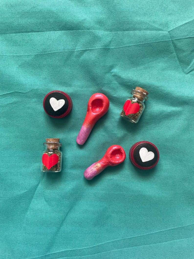 Pode incluir: Uma variedade de itens feitos &agrave; m&atilde;o, incluindo dois pequenos frascos de vidro com rolhas de corti&ccedil;a, cada um contendo um cora&ccedil;&atilde;o vermelho e ervas secas. Duas colheres vermelhas e rosa e dois c&iacute;rculos pretos e vermelhos com cora&ccedil;&otilde;es brancos.