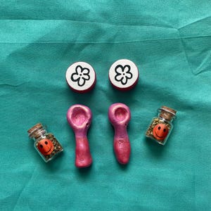 Pode incluir: Uma cole&ccedil;&atilde;o de pequenos itens feitos &agrave; m&atilde;o. Dois bot&otilde;es vermelhos e brancos com design de flores, dois cachimbos rosa e dois pequenos frascos de vidro com rolhas e adesivos de carinhas sorridentes. Os frascos cont&ecirc;m ervas verdes.