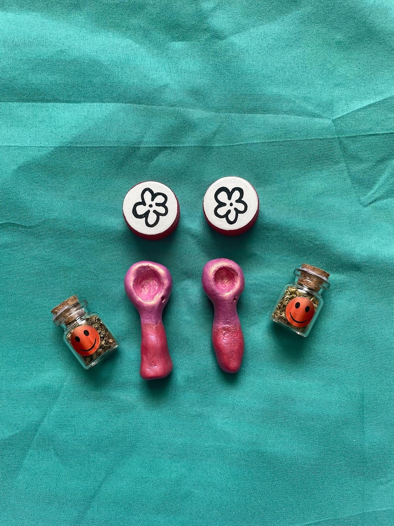Pode incluir: Uma cole&ccedil;&atilde;o de pequenos itens feitos &agrave; m&atilde;o. Dois bot&otilde;es vermelhos e brancos com design de flores, dois cachimbos rosa e dois pequenos frascos de vidro com rolhas e adesivos de carinhas sorridentes. Os frascos cont&ecirc;m ervas verdes.