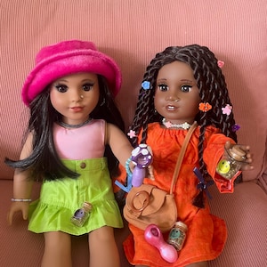 Pode incluir: Duas bonecas est&atilde;o sentadas em uma superf&iacute;cie de veludo cotel&ecirc; rosa. Uma boneca usa um chap&eacute;u rosa, um top rosa e uma saia verde. A outra boneca usa um vestido laranja e tem cabelos longos tran&ccedil;ados. Ambas as bonecas t&ecirc;m acess&oacute;rios.