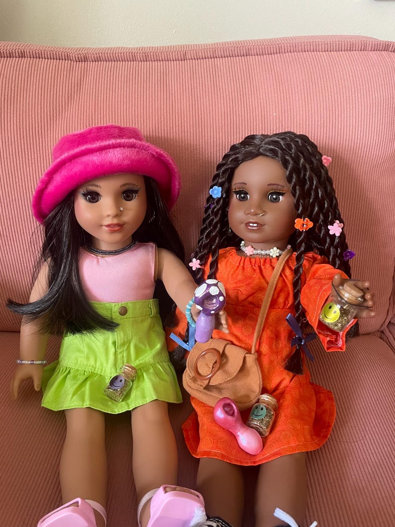 Pode incluir: Duas bonecas est&atilde;o sentadas em uma superf&iacute;cie de veludo cotel&ecirc; rosa. Uma boneca usa um chap&eacute;u rosa, um top rosa e uma saia verde. A outra boneca usa um vestido laranja e tem cabelos longos tran&ccedil;ados. Ambas as bonecas t&ecirc;m acess&oacute;rios.
